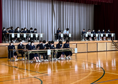 体育館のステージ上と手前に複数の長机と椅子が並べられ、生徒会役員のメンバーが席につき、生徒総会が行われている様子の写真