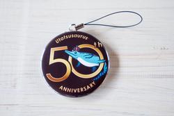 黒い背景に金色の『50th ANNIVERSARY』の文字と青い魚のキャラクターが描かれ、『Utaissaurus』の文字が入った缶バッジが黒いストラップに取り付けられている缶バッジストラップの写真