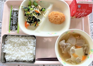 ピンクのトレーに給食が左から時計回りに味付けのり、小松菜の梅和え、かつおフライ、牛乳、油麩入りはっと汁、ごはんと並んでいる写真