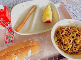 ピンクのトレーに給食が左から時計回りに牛乳、フランクフルト（パックケチャップ）、りんご、やきそば、コッペパンが並んでいる写真