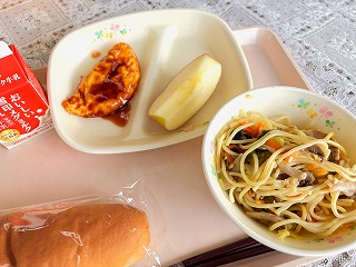 ピンクのトレーに給食が左から時計回りに牛乳、ミートオムレツ、入谷のりんご、和風きのこスパゲティー、米粉パンが並んでいる写真
