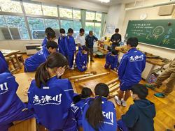 教室で体操服姿の生徒たちが、講師の男性と一緒に、床に並べた木材を囲んで指導を受けている写真