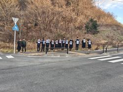 横断歩道がある道路わきの歩道で、草木が生えた斜面の前に横一列に並んだ生徒たちが、文字が書かれたプラカードを1枚ずつ持って立っている写真