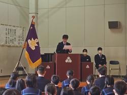 制服姿の生徒たちが整列して座る中、校章旗が飾られた舞台の演台に立った男子生徒が原稿を手に話をしている写真