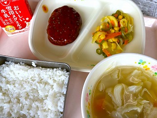 ピンクのトレーに給食が左から時計回りに牛乳、ハンバーグトマトソースがけ、マカロニのガーリックソテー、キャベツのコンソメスープ、ごはんと並んでいる写真