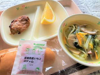 ピンクのトレーに給食が左から時計回りにタンドリーチキン、ジューシーフルーツ、きのこのスープスパゲティー、ソフトパンの上にいちごジャムが並べられている写真
