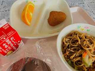 ピンクのトレーに給食が左から時計回りに牛乳、オレンジ、焼き栗コロッケ、やきそば、ココアパンと並んでいる写真