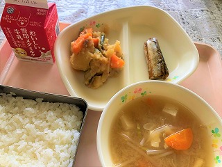 ピンクのトレーに給食が左から時計回りに牛乳、肉じゃが、さんまのおかか煮、もやしと油揚げのみそ汁、ごはんと並んでいる写真