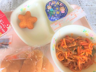 ピンクのトレーに給食が左から時計回りにパックソース、牛乳、星のコロッケ、七夕ゼリー、なすとトマトのスパゲティー、 バターロールと並んでいる写真