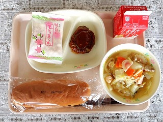 ピンクのトレーに給食が左から時計回りにひなあられ、手作りハンバーグ、牛乳、ちゃんぽんうどん、米粉パンが並んでいる写真