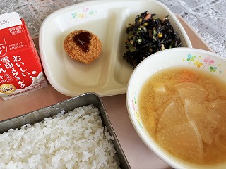 ピンクのトレーに給食が左から時計回りに牛乳、さけメンチ、ひじきと小松菜のごまマヨサラダ、大根と油あげのみそ汁、ごはんが並んでいる写真