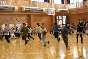体育館内で間隔をあけて並んだ数人の児童たちが体育館で元気よく飛び跳ねてダンスを踊っている写真