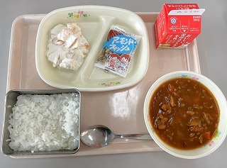 ピンクのトレーに給食が左から時計回りにフルーツのヨーグルトあえ、アーモンドフィッシュ、牛乳、ポークカレー、ごはんが並んでいる写真