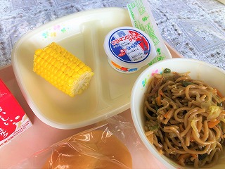 ピンクのトレーに給食が左から時計回りに牛乳、入谷産ゆでとうもろこし、ブラマンジェ（いちごソース）、やきそば、ミルクパンと並んでいる写真