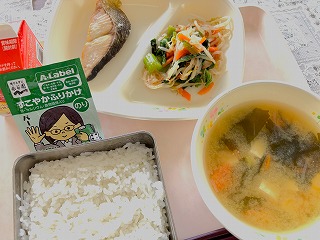 ピンクのトレーに給食が左から時計回りに牛乳、鮭の塩焼き、小松菜のおひたし、わかめ汁、ごはん、のりふりかけが並んでいる写真