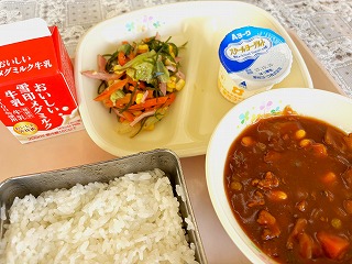 ピンクのトレーに給食が左から時計回りに牛乳、りっちゃんサラダ、ヨーグルト、ポークビーンズ、ごはんが並んでいる写真