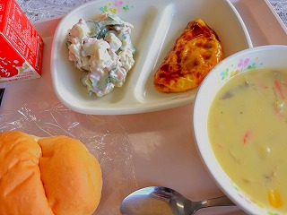 ピンクのトレーに給食が左から時計回りに牛乳、さつまいものサラダ、ミートオムレツ、コーンポタージュ、バターロールが並んでいる写真