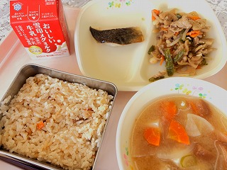 ピンクのトレーに給食が左から時計回りに牛乳、ほっけの塩焼き、レンコンと大豆のきんぴら、油ふのみそ汁、五目ごはんが並んでいる写真