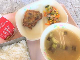 ピンクのトレーに給食が左から時計回りに牛乳、ポークピカタ、きゃべつと小松菜のソテー、なすと油ふのみそ汁、ごはんが並べられている写真
