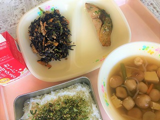 ピンクのトレーに給食が左から時計回りに牛乳、ひじきの煮物、あじの塩麹焼き、おくずかけ、ふりかけのかかったごはんと並んでいる写真
