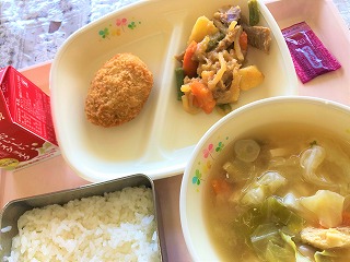 ピンクのトレーに給食が左から時計回りに牛乳、かつおかつ、肉じゃが、パックソース、しめじとキャベツのみそ汁、ごはんが並んでいる写真