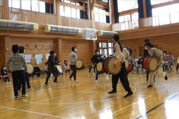 体育館の中で、円になった子どもたちが太鼓を胸にかけ、バチを持って「行山流水戸辺鹿子躍」の練習をしている写真