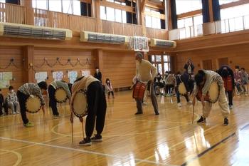 村岡さんが見守りながら、子どもたちが胸にかけた太鼓に体を覆いかぶせるように前かがみの姿勢をとって、両手には細長いバチを持ち、バチ先を体育館の床にまっすぐ下ろしてつけている様子の写真