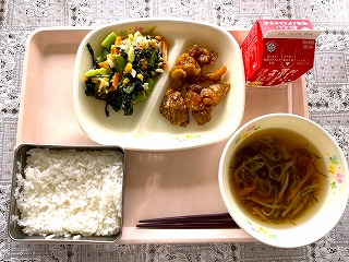 ピンクのトレーに給食が左から時計回りに小松菜とキャベツの和え物、チキンチキンごぼう、牛乳、もずくのスープ、ごはんと並んでいる写真