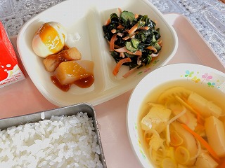 ピンクのトレーに給食が左から時計回りに牛乳、みそおでん、わかめときゅうりの梅あえ、すまし汁、ごはんが並んでいる写真