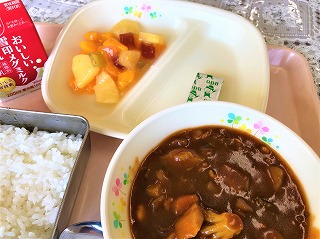 ピンクのトレーに給食が左から時計回りに牛乳、フルーツのゼリーあえ、チーズ、ポークビーンズ、ごはんが並べられている写真