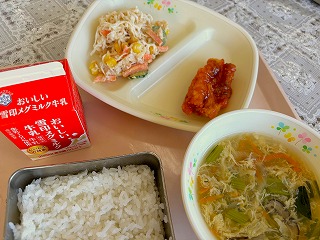 ピンクのトレーに給食が左から時計回りに牛乳、切干大根のかみかみサラダ、ヤンニョムチキン、玉子と小松菜の中華スープ、ごはんが並んでいる写真