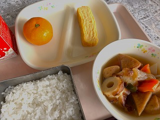 ピンクのトレーに給食が左から時計回りに牛乳、みかん、厚焼き玉子、煮込みおでん、ごはんが並んでいる写真
