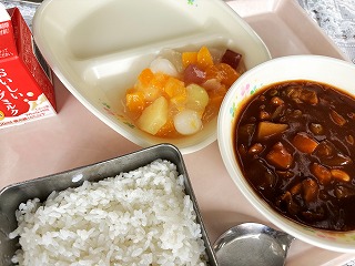 ピンクのトレーに給食が左から時計回りに牛乳、フルーツ白玉、ポークビーンズ、ごはんと並んでいる写真