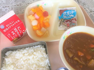 ピンクのトレーに給食が左から時計回りに牛乳、フルーツ白玉、アーモンドフィッシュ、ポークカレー、ごはんと並んでいる写真