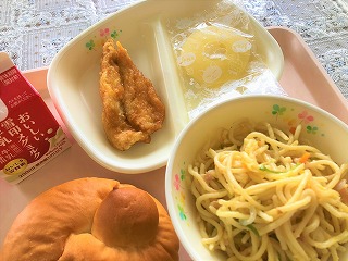 ピンクのトレーに給食が左から時計回りに牛乳、鶏肉のレモンソースがけ、冷凍パイ、春キャベツときのこのスパゲティー、ミルクパンが並んでいる写真