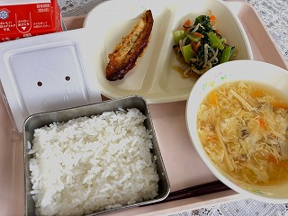 ピンクのトレーに給食が左から時計回りに牛乳、納豆、ささかまのみそマヨネーズ焼き、小松菜のごまあえ、かきたま汁、ごはんが並んでいる写真