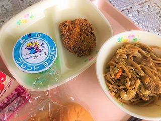 ピンクのトレーに給食が左から時計回りに牛乳、ヨーグルト、鶏肉の青のりフライ、焼きそば、ミルクパンと並んでいる写真