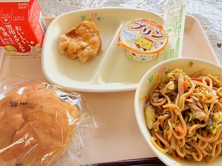 ピンクのトレーに給食が左から時計回りに牛乳、鶏肉のレモンソースがけ、豆乳プリン、やきそば、チョコチップパンが並んでいる写真