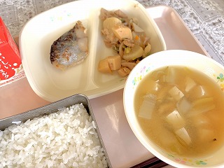 ピンクのトレーに給食が左から時計回りに牛乳、筑前煮、さわらの西京みそ焼き、めかぶと油あげのみそ汁、ごはんが並んでいる写真