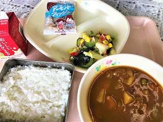 ピンクのトレーに給食が左から時計回りに牛乳、アーモンドフィッシュ、コーンとわかめのサラダ、ポークカレー、ごはんが並べられている写真