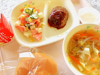 ピンクのトレーに給食が左から時計回りに牛乳、マセドアンサラダ、ハンバーグデミグラスソース、ワンタンスープ、豆乳プリン、ミルクパンが並んでいる写真