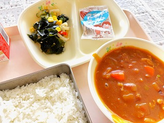 ピンクのトレーに給食が左から時計回りに牛乳、海藻サラダ、アーモンドフィッシュ、ポークカレー、ごはんが並んでいる写真