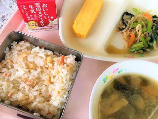 ピンクのトレーに給食が左から時計回りに牛乳、厚焼き卵、小松菜のおひたし、わかめのみそ汁、たけのこごはんと並んでいる写真