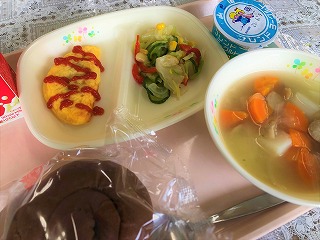 ピンクのトレーに給食が左から時計回りに牛乳、ミートオムレツ（パックケチャップ）、コーンサラダ、ヨーグルト、ポトフ、ココアパンが並んでいる写真