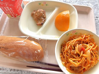 ピンクのトレーに給食が左から時計回りに牛乳、タンドリーチキン、いよかん、ナポリタン、米粉パンが並んでいる写真