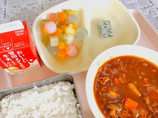 ピンクのトレーに給食が左から時計回りに牛乳、フルーツ白玉、チーズ、ポークビーンズ、ごはんが並んでいる写真
