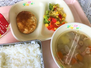 ピンクのトレーに給食が左から時計回りに牛乳、豆腐ハンバーグオニオンソース、ブロッコリーとコーンのソテー、ひよこ豆のスープ、ごはんが並べられている写真