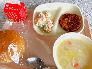 ピンクのトレーに給食が左から時計回りに牛乳、入谷産じゃがいものポテトサラダ、ハムカツ（ソースがけ）、コロコロ野菜の豆乳スープ、丸パンが並んでいる写真