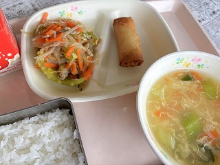 ピンクのトレーに給食が左から時計回りに牛乳、野菜たっぷりチャプチェ、春巻き、中華コーンスープ、ごはんが並んでいる写真