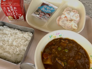 ピンクのトレーに給食が左から時計回りに牛乳、アーモンドフィッシュ、フルーツのヨーグルトあえ、ポークカレー、ごはんと並んでいる写真